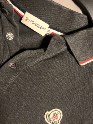 Mörkgrå Moncler pikétröja - Snygg mörkgrå långärmad pikétröja från Moncler med klassisk krage och knappar framtill. Står storlek XXL, men passar som M! kom privat för mer information! 