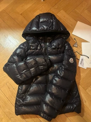 Svart dunjacka från Moncler med luva - Snygg svart dunjacka från Moncler med luva och dragkedja framtill. Jackan har två sidofickor med dragkedja och Moncler-logga på ärmen. Insidan har ett unikt serietryck. Perfekt för kalla dagar. Storleken står med siffrorna 12 och 152 cm, men passar mig som är 165 (lite kort men passar ganska bra)