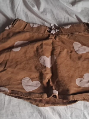Brun kjol med hjärtmönster storlek 98 till 104 cm - Söta bruna kjol i mjuk bomull med ljusa hjärtan som mönster. De har en midja med knytband och kort, loose passform. Perfekta för varma sommardagar eller som mysplagg hemma.