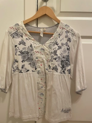 Vit blommig blus från Honey Ocean Earth - Surfmärke Nyskick, använd en gång. Mjuk och skön. vit blus med svarta blommönster och diskreta röda detaljer. Blusen har trekvartsärm och v-ringning med knappar framtill. Materialet känns mjukt och lätt, perfekt för en avslappnad look. Märket Honey ocean earth är broderat längst ner på framsidan. Passar small och medium 