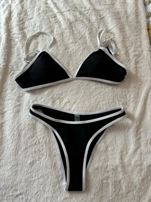 Svart bikini med vita kanter - Snygg svart bikini med vita kontrastkanter. Överdelen har trekantskupor och smala axelband, medan underdelen har en klassisk, något högre skärning. Perfekt för stranden eller poolen och ger en stilren look.