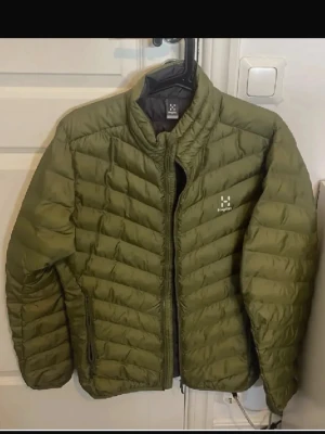 Olivgrön dunjacka från Haglöfs - Snygg olivgrön dunjacka från Haglöfs med quiltad design och hög krage. Jackan har dragkedja framtill, två sidofickor med dragkedja och diskret Haglöfs-logga på bröstet. Perfekt för kalla vinterdagar och riktigt lätt att bära.