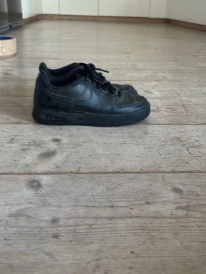 Svarta Nike Air Force 1 sneakers - Klassiska helsvarta Nike Air Force 1 sneakers i läder med svart sula och svarta skosnören. Modellen har en låg siluett och diskret Nike-logga på sidan. Perfekta för dig som gillar en clean och tidlös streetstil.