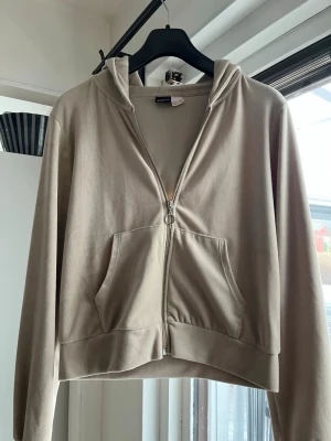 Beige zip hoodie från Gina Tricot - Snygg beige hoodie från Gina Tricot med dragkedja framtill och stor känguruficka. Hoodien har huva och långa ärmar, perfekt för en chill och avslappnad stil. Tillverkad i mjukt bomullsliknande material.