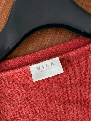 Rostfärgad kofta från Vila - Mysig öppen kofta från Vila i en snygg rostfärg. Koftan har långa ärmar, två stora fickor framtill och ribbade muddar vid ärmslut och nederkant. Perfekt att slänga över en t-shirt för en avslappnad look.