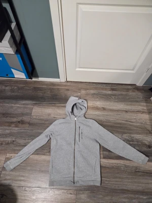 Grå hoodie från Sail Racing - Snygg grå hoodie från Sail Racing med dragkedja hela vägen och en extra bröstficka med dragkedja. Klassisk huva och diskret logga på bröstet. Perfekt för en avslappnad och sportig stil. Mjuk bomullsblandning för skön känsla.