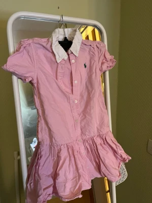 Rosa skjortklänning från Ralph Lauren till barn  - Söt rosa barn skjortklänning från Ralph Lauren med vit krage och korta puffärmar. Klänningen har knappar framtill, volang i nederkant och den klassiska loggan broderad på bröstet. Perfekt för en fräsch och avslappnad look.