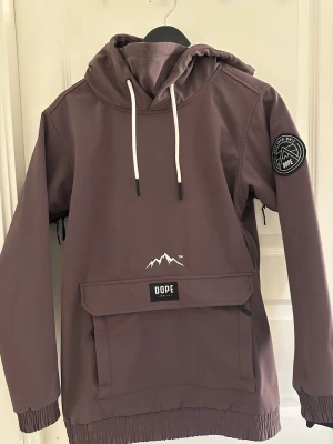 Lila hoodie från Dope med huva - Snygg lila vindjacka från Dope med huva och stor magficka framtill. Jackan har vita snören i huvan, patch på ärmen och dragkedjor i sidorna. Perfekt för dig som gillar streetwear och outdoor-vibe. Har Tyväär växt ur denna jacka så den har inte kommit till användning.