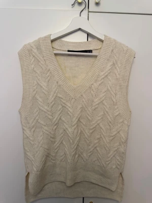 Beige stickad väst med v-ringning - Snygg beige/vit stickad väst med v-ringad hals och grovt kabelstickat mönster. Perfekt att slänga över en skjorta eller t-shirt för en trendig lager-på-lager-look. Ribbad kant nedtill och vid halsen för extra stilpoäng.
