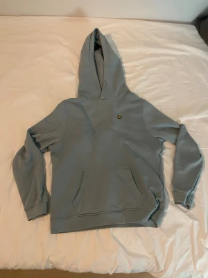 Ljusblå hoodie från Lyle & Scott - Snygg ljusblå hoodie från Lyle & Scott med klassisk gul fågel-logga på bröstet. Hoodien har stor huva, magficka och ribbade muddar vid ärmar och nederkant. Perfekt för en chill och avslappnad stil. Mjuk bomullskänsla och enkel att matcha med jeans eller joggers. 40 cm i axlarna och 60 i längd 