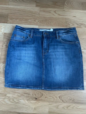 Blå jeanskjol med klassisk passform - Snygg blå jeanskjol i klassisk femficksmodell med bälteshällor och knappstängning framtill. Kjolen har en rak, kort passform och är tillverkad i denim. Perfekt för dig som gillar en enkel och tidlös stil. Kan sänka priset!!