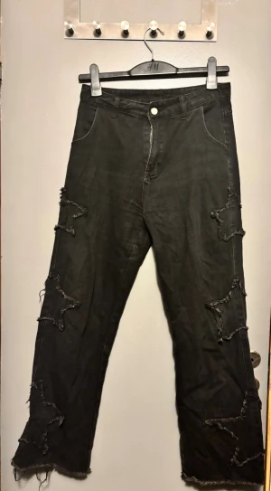 Svarta jeans med stjärn-detaljer Shein - Säljer ett par svarta jeans från Shein med unika stjärnformade slitningar längs benen och råa kanter vid bensluten. Jeansen har klassisk fick modell och knappgylf. Perfekta för dig som gillar en edgy och personlig stil.
