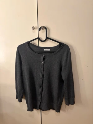 Mörkgrå kofta från Jacqueline de Yong - Stilren mörkgrå kofta från Jacqueline de Yong med rund halsringning och knäppning framtill. Koftan har trekvartsärmar och är tillverkad i ett mjukt, tunt stickat material som passar perfekt till lager på lager.