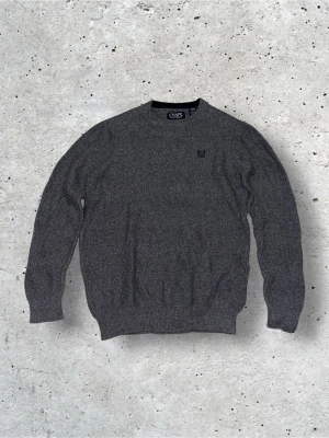 Stickad Chaps tröja - Grå stickad tröja från Chaps med rund halsringning och diskret logga broderad på bröstet. Tröjan har ribbade muddar vid ärmslut och nederkant, vilket ger en klassisk och stilren look. Perfekt för lager på lager under kyliga dagar. Storlek S.