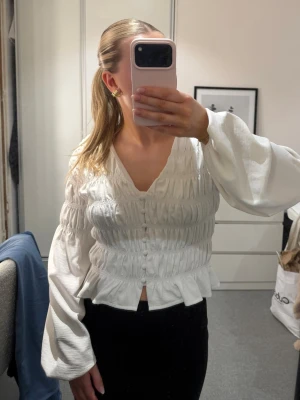 Vit blus  - Säljer en vit blus från Gina Tricot i storlek XS. Blusen har djup v-ringning, knappar framtill, smockad midja och volang nedtill🤍Använd en gång