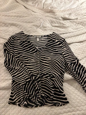 Zebramönstrad blus från H&M - Snygg zebramönstrad blus från H&M med svartvita ränder och v-ringning. Blusen har långa ärmar och knappar framtill. Perfekt för dig som vill sticka ut med ett trendigt djurmönster. Tunn och luftig i materialet.