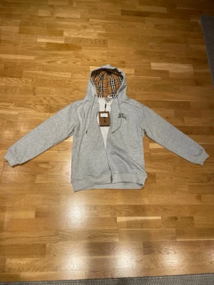 Grå zip hoodie från Burberry - Snygg grå zip hoodie från Burberry med klassiskt rutigt foder i huvan och broderad logga på bröstet. Tillverkad i mjuk bomullsblandning och har dragsko i huvan samt ribbade muddar. Perfekt för en avslappnad men stilren look.