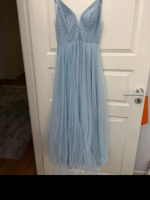 Ljusblå långklänning med pärlor - Superfin ljusblå långklänning med glittrig tyllkjol och dekorerad överdel med pärlor. Klänningen har smala axelband och en v-ringad urringning. Perfekt för bal eller festliga tillfällen. Materialet är troligtvis polyester och tyll.