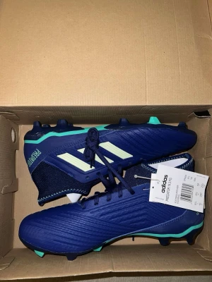 Adidas Predator blå fotbollsskor - Snygga Adidas Predator  fotbollsskor i blått med vita och turkosa detaljer. Skorna har klassisk snörning, stickad strumpliknande krage och tålig syntetisk ovandel. Perfekta för dig som vill ha helt nya fotbollsskor,skorna har inte använts och kan både ha till konstgräs och vanlig gräs.