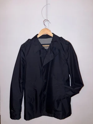 Herno overshirt/rock - Snygg mörkblå rock/ocershirt med klassisk krage och knäppning framtill. Jackan har raka linjer, axelklaffar och diskreta sidfickor. Perfekt för dig som gillar stilren och tidlös design. Passar till både skjorta och t-shirt. Den har en defekt med en söndrig knapp på ena ärmen. 