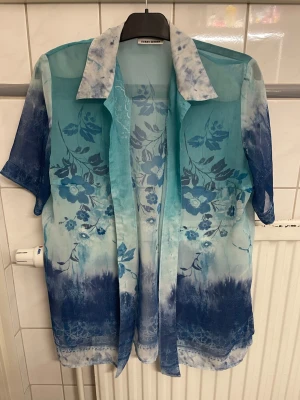 Blå transparent blommig skjorta - Säljer en blå transparent skjorta från Gerry Weber med korta ärmar och blommigt mönster. Skjortan har en avslappnad passform, klassisk krage och är tillverkad i ett tunt syntetmaterial. Perfekt för sommarens varma dagar!