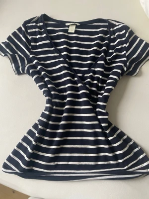 Randig omlottopp från H&M Mama - Mörkblå och vit randig omlottopp från H&M Mama. Toppen har korta ärmar, v-ringning och är tillverkad i mjuk bomull. Perfekt för en avslappnad och bekväm stil.