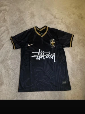 Svart Brasilien x Stüssy fotbollströja - Snygg svart fotbollströja från Nike i samarbete med Stüssy och Brasilien. Tröjan har gulddetaljer på krage och ärmslut, Brasilien-emblem på bröstet och Stüssy-logga i vitt framtill. Perfekt för dig som gillar streetwear och fotboll.
