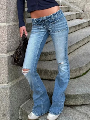 Ljusblå bootcut jeans med snörning - Säljer ett par ljusblå bootcut jeans med snörning i midjan och slitningar vid knäna. Jeansen har en låg midja och coola fickdetaljer framtill. Perfekta för dig som gillar en trendig och avslappnad stil.