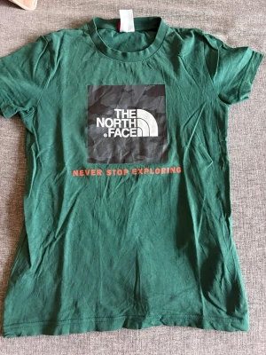 Grön t-shirt från The North Face - Snygg grön t-shirt från The North Face med klassisk logga och texten 'Never Stop Exploring' i rött framtill. T-shirten har rund hals och korta ärmar, tillverkad i mjuk bomull som är skön att bära hela dagen.