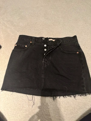 Svart jeanskjol från Levi's - Säljer en svart jeanskjol från Levi's med rå kant nertill och knappgylf framtill. Kjolen har klassiska fem fickor och en cool, lite sliten look. 