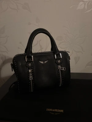 Svart handväska Zadig & Voltaire - Snygg svart handväska från Zadig & Voltaire i strukturerat läder. Väskan har två korta handtag, silverfärgade detaljer, dubbla dragkedjor framtill och dekorativa nitar längs sidorna. Perfekt för dig som gillar en rockig och stilren look.