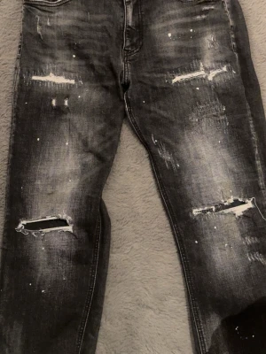 Svarta slitna jeans med hål - Säljer ett par svarta jeans med slitningar, hål och vit färgdetalj. Jeansen har en tvättad look och raka ben. Perfekta för en streetwear-stil och passar dig som gillar en edgy vibe.