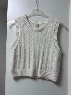 Vit stickad väst från Lindex - Snygg vit stickad väst från Lindex med klassiskt kabelstickat mönster. Croppad modell med bred ribbad kant nertill och vid ärmöppningarna. Perfekt att slänga över en skjorta eller t-shirt för en trendig lager-på-lager-look.