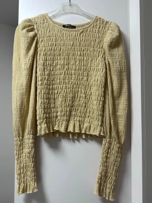 Gul smockad långärmad topp - ginatricot - Trendig gul topp med smockat tyg och långa ärmar. Modellen har rund halsringning och lätt puffade axlar för en cool siluett. Perfekt att styla med jeans eller kjol för en fräsch look.