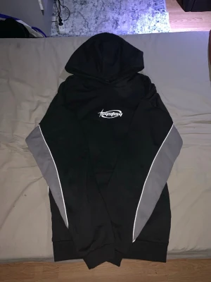 Svart hoodie från System med grå detaljer - Svart hoodie från System med grå paneler längs ärmarna och vita linjer. Tryckt vit logga på bröstet. Klassisk huva och ribbade muddar. Perfekt för en streetwear-look.