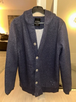 Hoodie Cardigan - Mysig mörkblå hoodiekofta från Ombre med knappar framtill och huva. Tillverkad i mjukt sweatshirtmaterial, perfekt för chill dagar. Långärmad modell med ribbade muddar och enkel, stilren design.