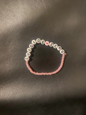 Pärlarmband med text och rosa hjärta - Sött armband med vita bokstavspärlor som stavar 'SUICIDEZERO' och ett rosa hjärta. Resten av armbandet består av små ljusrosa pärlor. Perfekt accessoar för dig som gillar personliga smycken och vill bära ett statement.