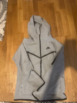 Nike tech fleece  - Snygg grå hoodie från Nike med svart dragkedja och svarta detaljer längs sömmarna. Klassisk huva och Nike-logga på bröstet. Tillverkad i mjukt material som känns skönt mot huden. Perfekt för en avslappnad och sportig stil.