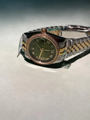 Rolx Datejust grön/guld klocka - Exklusiv Rolx Oyster Perpetual Datejust med grön urtavla, romerska siffror och gulddetaljer. Boetten är dekorerad med gnistrande stenar och armbandet är tvåfärgat i silver och guld. Perfekt för dig som gillar lyxiga accessoarer. Boettdiameter 36 mm, mekaniskt urverk, lasergraverad markering vid klockan 6, logotyp stämplad i de fyra hörnen på baksidan.✅