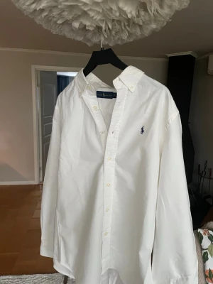 Vit skjorta från Ralph Lauren - Klassisk vit skjorta från Ralph Lauren med blå broderad logga på bröstet. Skjortan har knappar framtill, krage och långa ärmar. Tillverkad i mjuk bomull för en clean och tidlös look.