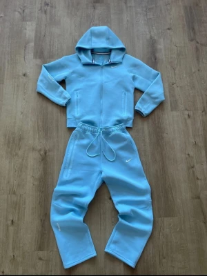 Ljusblå hoodie från Nike NOCTA - Snygg ljusblå hoodie från Nike NOCTA med dragkedja och huva. Hoodien har minimalistisk design med små vita loggor och är gjord i mjukt material som känns riktigt skönt mot huden. Perfekt för en chill och sportig stil.