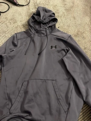 Grå hoodie från Under Armour - Snygg grå hoodie från Under Armour med svart logga på bröstet. Hoodien har huva med dragsko och en stor magficka framtill. Tillverkad i mjukt material som passar perfekt för en avslappnad stil.