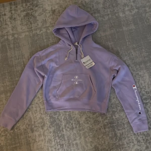 Ljuslila hoodie från Champion - Säljer en jättesnygg ljuslila hoodie från Champion med en ficka där fram, broderad logga framtill och en logga på ärmen samt vit dragsko i huvan. Hoodien har en halv dragkedja framtill och är i mjukt bomullsmaterial. Perfekt för en avslappnad och sportig stil. Hoodien är helt ny med prislapp på. 