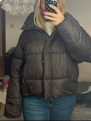 Svart pufferjacka med hög krage - Säljer en svart pufferjacka med hög krage och breda kanaler. Jackan har dragkedja och tryckknappar framtill samt en boxig, oversized passform. Perfekt för kalla vinterdagar och riktigt skön att bära. Passar dig som gillar en stilren och enkel look.