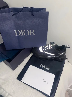 Svarta Dior sneakers med vita detaljer - Snygga svarta Dior sneakers med vita reflekterande detaljer och chunky sula. Skon har en modern design med mesh och lädermaterial, samt tydlig Dior-logga på sidan. Perfekta för dig som vill ha en exklusiv och trendig look.