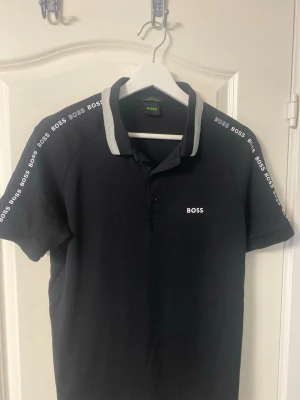 Svart pikétröja från BOSS - Snygg svart pikétröja från BOSS med vit logga på bröstet och längs axlarna. Kragen har en grå kant och tröjan har knappar framtill. Perfekt för en clean och sportig look.