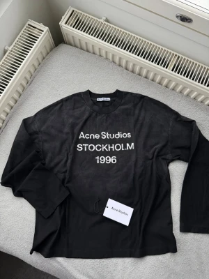Svart långärmad  skjorta Acne Studios - Cool svart långärmad skjorta från Acne Studios med vit text 'Acne Studios STOCKHOLM 1996' på bröstet. Klassisk rund halsringning och relaxed fit. Perfekt för dig som gillar minimalistisk och stilren design.