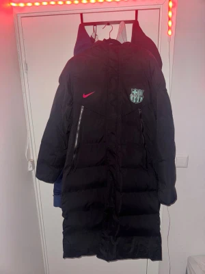 Svart lång dunjacka FC Barcelona Nike - Säljer en svart lång dunjacka från Nike med FC Barcelona-logga på bröstet. Jackan har huva, två snedställda dragkedjefickor framtill och en röd Nike-logga. Perfekt för kalla vinterdagar och riktigt snygg streetstil. Size xl sitter som L/M