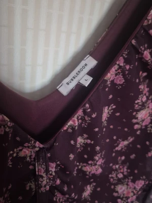 Blommig vinröd klänning Bubbleroom - Söt vinröd kortklänning från Bubbleroom med rosa blommönster, v-ringning och trekvartsärm. Klänningen har rynkade detaljer framtill och volangkant nedtill. Perfekt för dig som gillar romantisk stil och vill sticka ut lite extra.