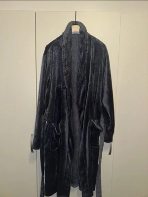 Mörkgrå fluffig morgonrock - Mysig och fluffig morgonrock i mörkgrå färg med två stora fickor framtill och knytskärp i midjan. Tillverkad i mjukt, lent material som känns skönt mot huden. Perfekt för chilliga morgnar eller sena kvällar hemma.